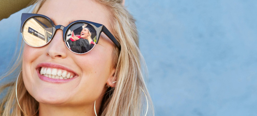 Woman smiling wearing sunglasses reflecting UHI Inverness campus building and graduates