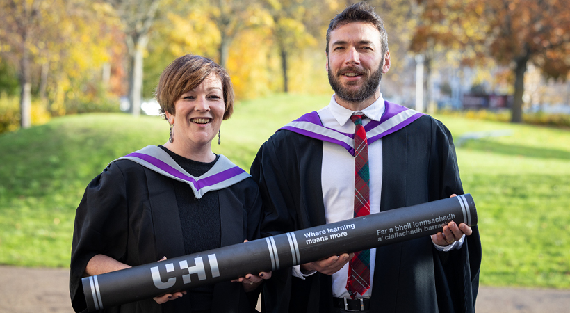 Two UHI Inverness Graduates wearing graduating robes and holding an oversized Graduation scroll Two UHI Inverness Graduates wearing graduating robes and holding an oversized Graduation scroll